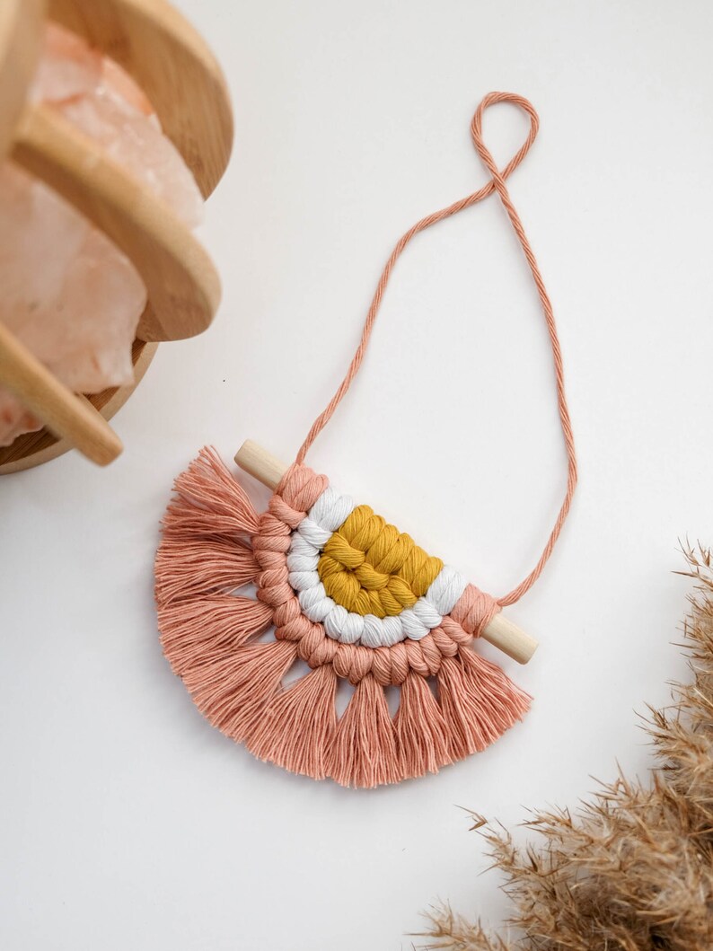 May include: A pink and yellow macrame wall hanging with a wooden dowel and fringe. The hanging is attached to a string.