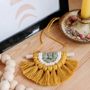 May include: A small, handmade macrame wall hanging with a yellow, white, and green color scheme. The hanging features a half-circle shape with a fringe border.