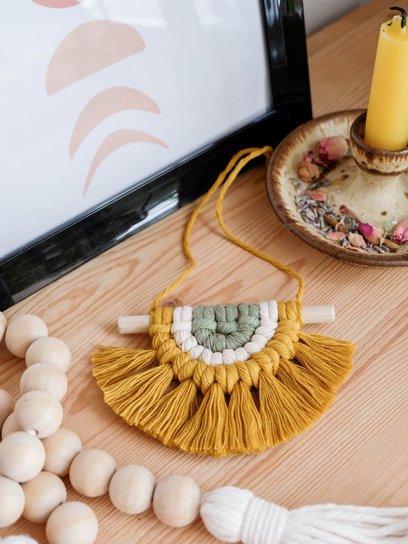 May include: A small, handmade macrame wall hanging with a yellow, white, and green color scheme. The hanging features a half-circle shape with a fringe border.
