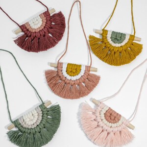 May include: Five colorful macrame wall hangings with wooden dowels and fringe. Each hanging features a different color combination of pink, green, yellow, and white.