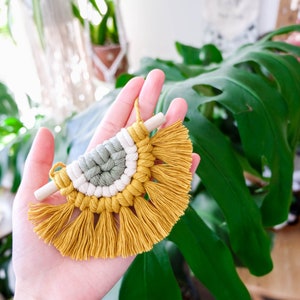 May include: A small, half-circle macrame wall hanging with a wooden dowel at the top. The macrame is made with yellow, white, and green yarn and has a fringe at the bottom.