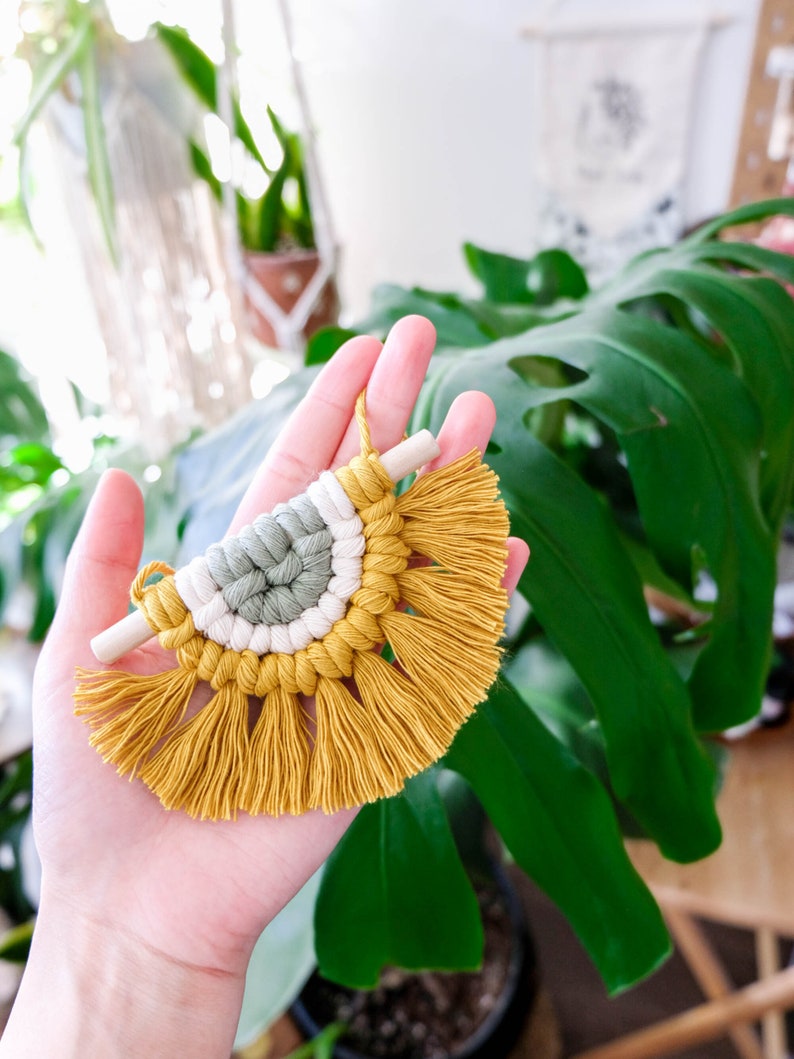 May include: A small, half-circle macrame wall hanging with a wooden dowel at the top. The macrame is made with yellow, white, and green yarn and has a fringe at the bottom.