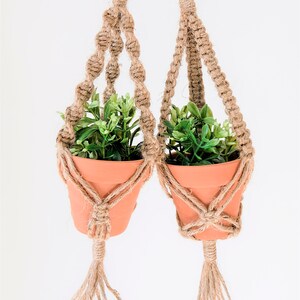 May include: Two terracotta pots containing green plants are suspended from macrame hangers with metal rings. The hangers are made of natural jute rope with tassels.