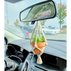 Op de afbeelding: Een kleine plant in een pot die aan de achteruitkijkspiegel van een auto hangt. De plant zit in een terracotta pot en is omgeven door een macramé hanger.