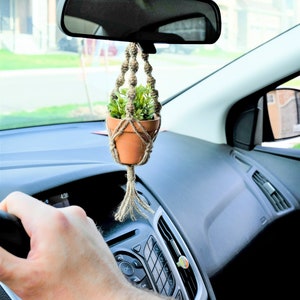 May include: A small potted plant hanging from the rear view mirror of a car. The plant is in a terracotta pot and is surrounded by a macrame hanger. The plant is green and the hanger is beige.
