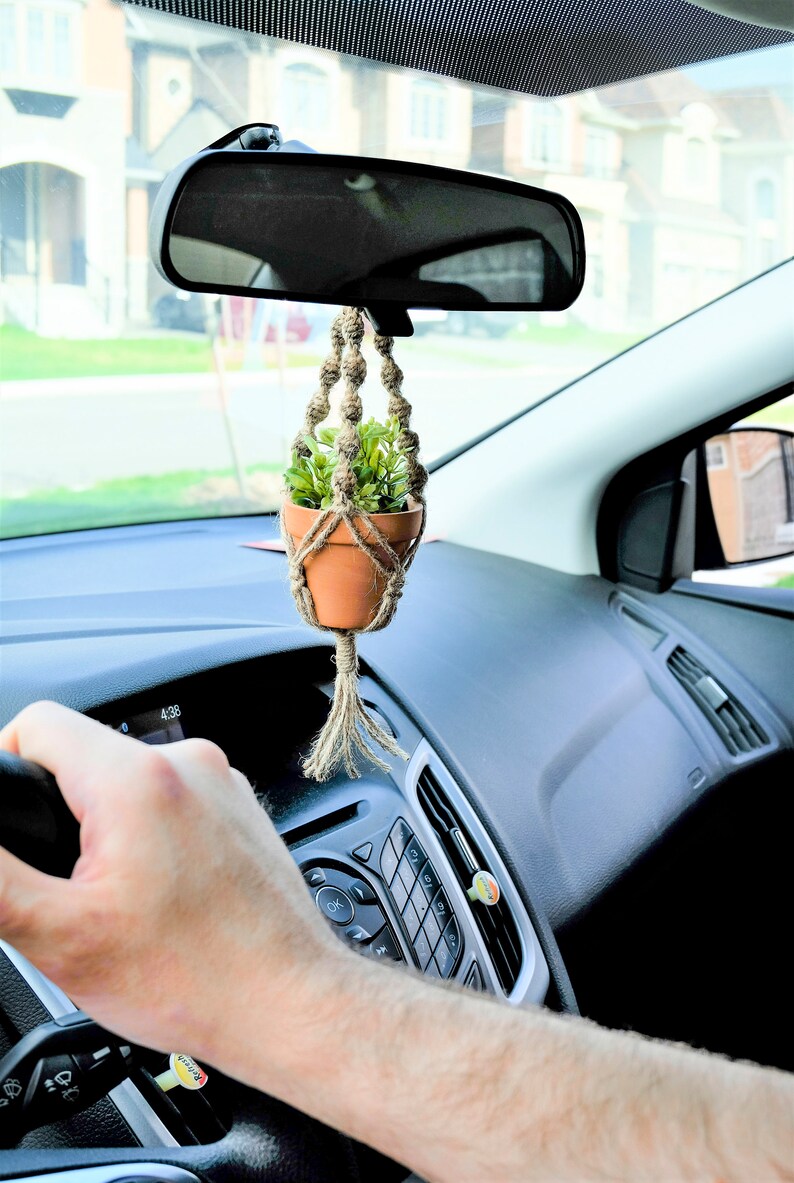 May include: A small potted plant hanging from the rear view mirror of a car. The plant is in a terracotta pot and is surrounded by a macrame hanger. The plant is green and the hanger is beige.
