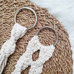 May include: Two white macrame plant hangers with silver metal rings. The hangers are on a woven brown background.
