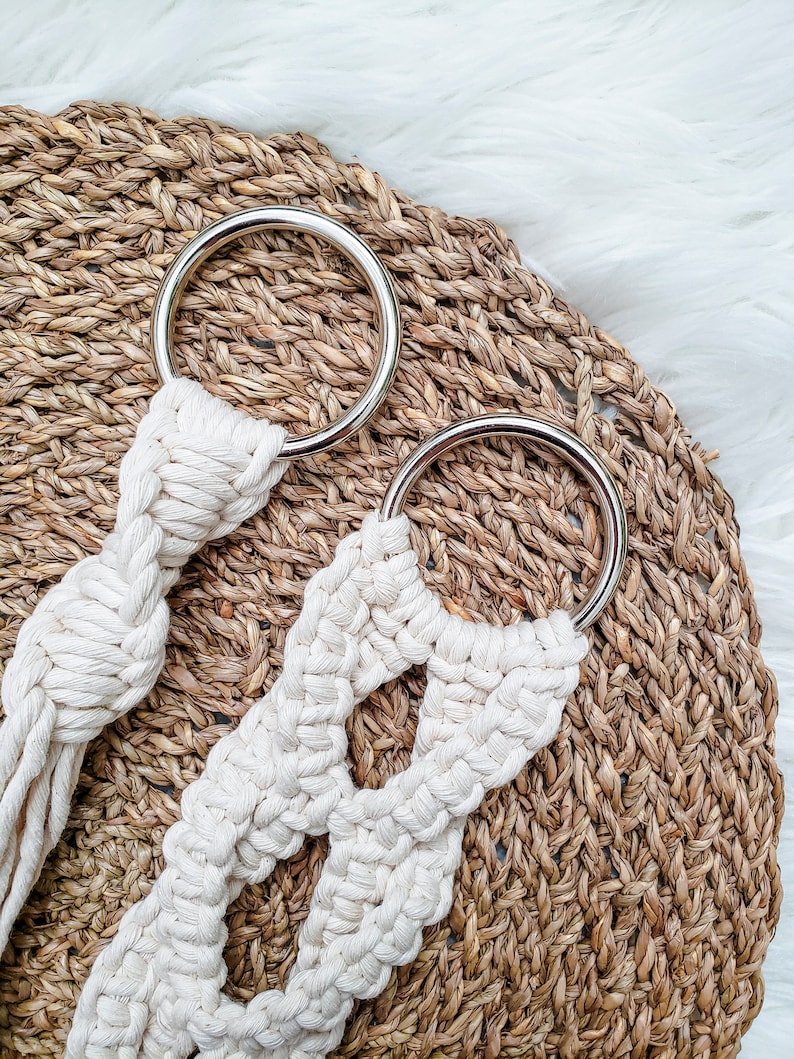 May include: Two white macrame plant hangers with silver metal rings. The hangers are on a woven brown background.