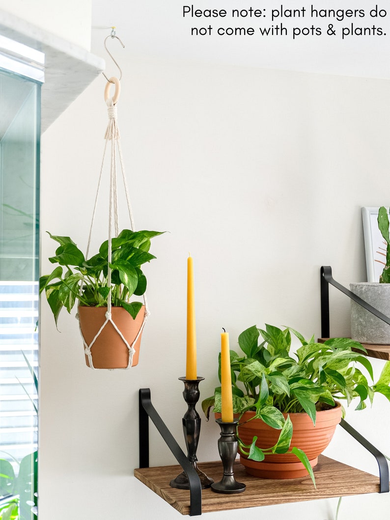 May include: A white macrame plant hanger with a wooden ring hangs from a hook on a white wall. A terracotta pot with a green plant is hanging from the macrame hanger. Two candlesticks with yellow candles are on a wooden shelf with black metal brackets. The shelf is attached to the white wall. The text "Please note: plant hangers do not come with pots & plants." is written on the wall above the hanger.