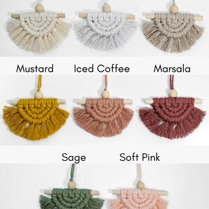 May include: Nine macrame wall hangings in various colors, including natural, blue thistle, oatmeal, mustard, iced coffee, marsala, sage, and soft pink. Each hanging features a wooden dowel and a wooden bead.