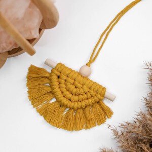 May include: A mustard yellow macrame wall hanging with a wooden dowel and a wooden bead. The macrame is in the shape of a half circle with fringe on the bottom.