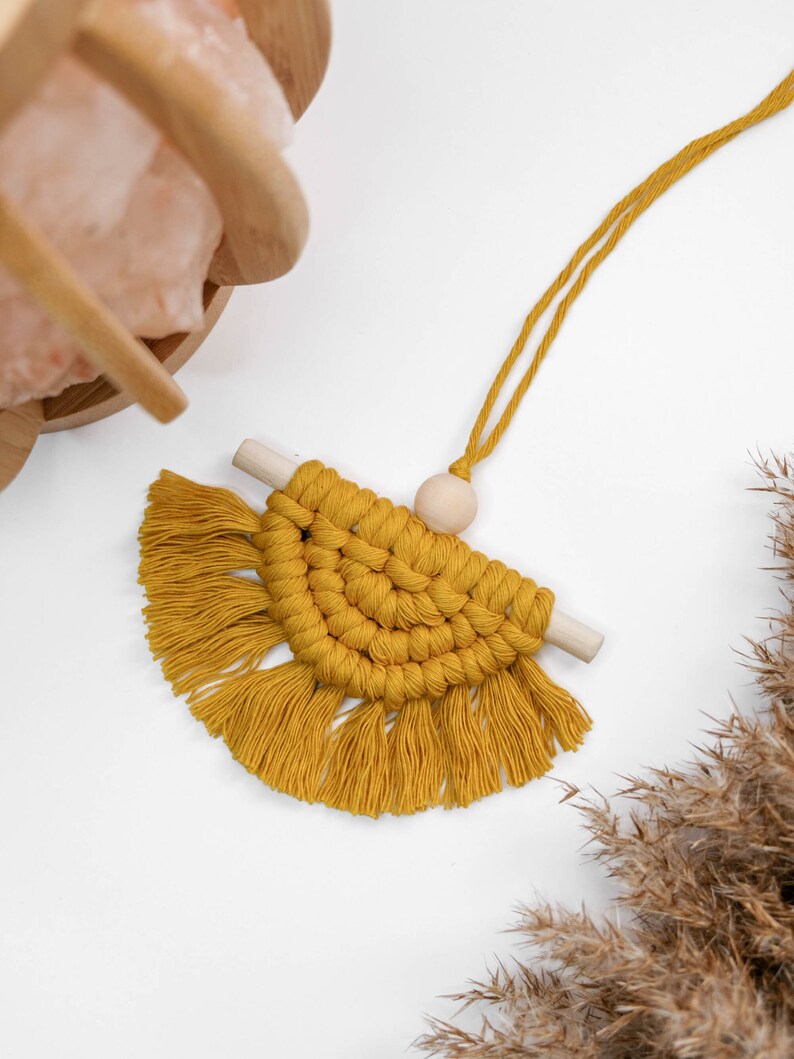 May include: A mustard yellow macrame wall hanging with a wooden dowel and a wooden bead. The macrame is in the shape of a half circle with fringe on the bottom.