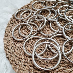 May include: A close-up of a pile of silver metal rings on a woven brown surface. The rings are all the same size and shape, and they are arranged in a random pattern.