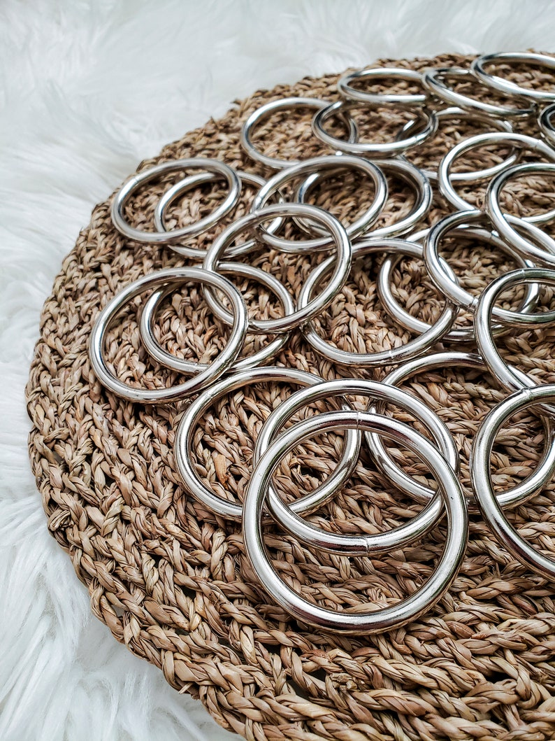 May include: A close-up of a pile of silver metal rings on a woven brown surface. The rings are all the same size and shape, and they are arranged in a random pattern.