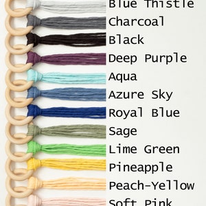 May include: A set of 14 wooden rings with different colored yarn tassels attached. The colors include natural cotton, sand, blue thistle, charcoal, black, deep purple, aqua, azure sky, royal blue, sage, lime green, pineapple, peach-yellow, soft pink, iced coffee, and marsala.