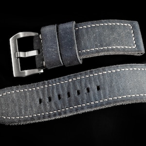 May include: Close-up of a gray watch strap with white stitching. The strap has a silver buckle and is made of a textured material. The brand name "THOMAS ARTISAN STRAPS" is in the top left corner.
