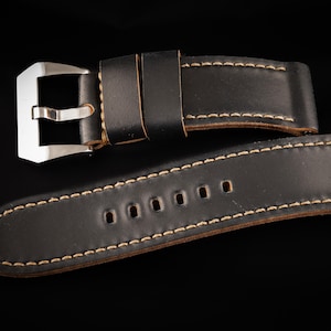 May include: Black leather watch strap with a contrasting light brown stitching. The strap has a silver buckle and five holes for adjusting the fit.