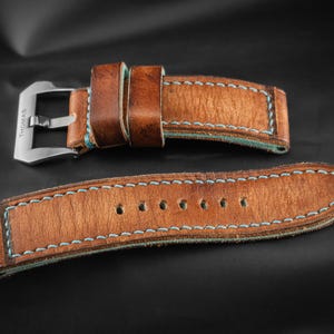 Puede incluir: Correa de reloj de cuero marrón de dos piezas con costuras azul claro. La correa tiene una hebilla plateada y varios agujeros para ajustar. El cuero tiene un aspecto envejecido. La marca "THOMAS ARTISAN STRAPS" es visible.