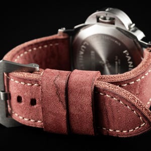 May include: Close-up of a watch with a reddish-brown leather strap. The strap has white stitching and a silver buckle. The watch face is visible, with the brand name "PANERAI" and other text. The strap is made by THOMAS ARTISAN STRAPS.