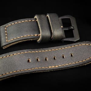 Könnte beinhalten: Nahaufnahme eines schwarzen Leder-Uhrenarmbands mit beigen Nähten. Das Armband hat eine schwarze Metallschnalle und mehrere Löcher zur Anpassung. Das Leder wirkt abgenutzt und verleiht ihm einen Vintage-Look. Das Armband ist für eine Uhr konzipiert.