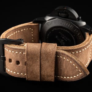 May include: Close-up of a brown leather watch strap with white stitching and a black buckle. The watch strap is attached to a black watch face. The brand name "THOMAS ARTISAN STRAPS" is visible in the upper left corner.