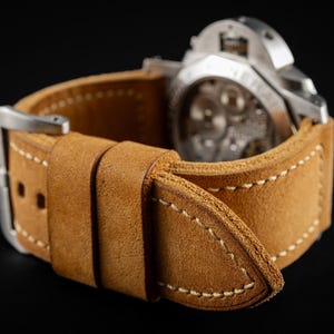 May include: Close-up of a tan leather watch strap with white stitching and a silver buckle. The watch face is visible, showcasing the intricate internal mechanisms. The strap is designed to fit a large watch.
