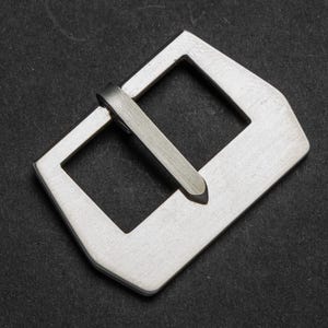 Puede incluir: Hebilla de reloj plateada con una abertura rectangular y un pasador. La hebilla tiene un acabado de metal cepillado y está sobre un fondo oscuro. La marca "THOMAS ARTISAN STRAPS" está en la esquina superior izquierda.