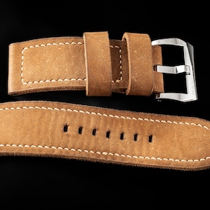 May include: A light brown leather watch strap with white stitching and a silver buckle. The strap is made of suede-like material and has several holes for adjustment. The brand name "THOMAS ARTISAN STRAPS" is visible in the top left corner.