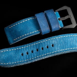 May include: A close-up of a blue leather watch strap with white stitching. The strap has a silver buckle and is set against a black background. The brand name "THOMAS ARTISAN STRAPS" is visible in the top left corner.