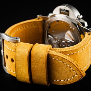 May include: Close-up of a watch with a yellow-tan leather strap. The strap is stitched with white thread and has a silver buckle. The watch face is visible, showing the intricate internal mechanisms.