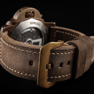 May include: Close-up of a brown leather watch strap with a bronze buckle. The strap has white stitching and a suede-like texture. The watch case is visible in the background, with the brand name "THOMAS ARTISAN STRAPS" in the top left corner.