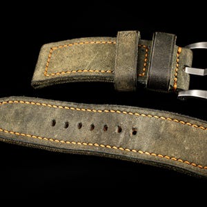 May include: A close-up of a watch strap made of olive green suede leather with orange stitching. The strap has a silver buckle and is set against a black background. The brand name "THOMAS ARTISAN STRAPS" is visible in the top left corner.