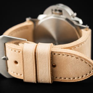 May include: Close-up of a light beige leather watch strap with a silver buckle. The strap is stitched with a contrasting thread and is attached to a silver watch face. The brand name "THOMAS ARTISAN STRAPS" is visible in the top left corner.