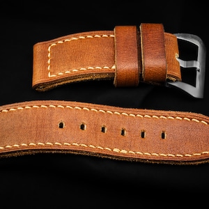 Könnte beinhalten: Nahaufnahme eines braunen Leder-Uhrenarmbands mit cremefarbenen Nähten. Das Armband hat eine silberne Schnalle und ist vor einem schwarzen Hintergrund platziert. Das Armband ist aus hochwertigem Leder gefertigt.