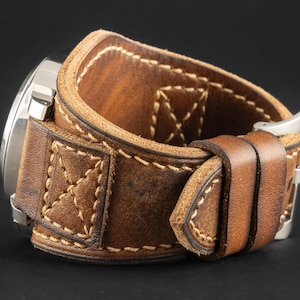 Può includere: Cinturino per orologio in pelle marrone con fibbia in acciaio inossidabile. Il cinturino è spesso e presenta cuciture decorative. Il quadrante dell'orologio è visibile sul lato sinistro dell'immagine. Il cinturino è progettato per un polso grande.