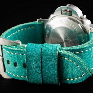 May include: Close-up of a teal leather watch strap with cream stitching and a silver buckle. The watch face is visible, with the text "OFFICINE PANERAI" on the back. The strap is made by THOMAS ARTISAN STRAPS.