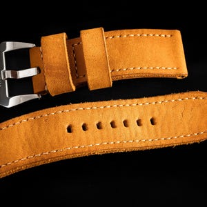 May include: Close-up of a tan leather watch strap with white stitching. The strap has a silver buckle with the word "THOMAS" engraved on it. The strap is made of a soft, suede-like material.