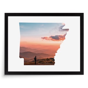 May include: Framed print featuring the outline of Arkansas filled with a scenic landscape. The image shows a person standing on a hilltop, silhouetted against a sunset with orange and blue hues. The frame is black.