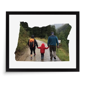 May include: Framed print featuring the outline of Oregon, filled with a family walking on a path. The family includes a person wearing an orange backpack, a child in a red jacket, and a person with hiking poles. The image is set against a white background.