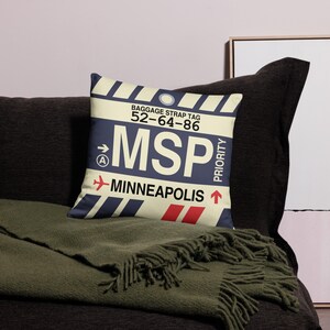 May include: A decorative cushion featuring a vintage luggage tag design. The tag is yellow with black stripes and displays the text "BAGGAGE STRAP TAG 52-64-86 MSP MINNEAPOLIS" in blue and red. The tag also includes a red arrow pointing right and a red arrow pointing upwards with the word "PRIORITY" written vertically.