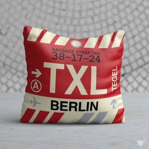 May include: Red and white cushion with a luggage tag design. The tag says "Baggage Strap Tag 38-17-24 TXL Berlin Tegel".