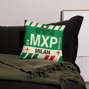 May include: A cushion with a green and white baggage tag design. The text "MXP MILAN" is displayed in white, along with the airport code and other details. The cushion is placed on a dark grey surface.