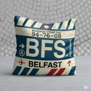 May include: A white pillow with a blue and red luggage tag design. The tag says "Baggage Strap Tag 94-76-08 BFS BELFAST" with an arrow pointing to the right and a small airplane icon. The tag also says "Aldergrove" with an arrow pointing upwards.