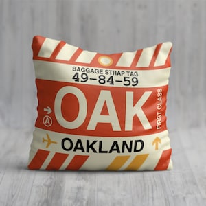 May include: A red, white and yellow throw pillow with a luggage tag design. The tag says "Baggage Strap Tag 49-84-59 OAK OAKLAND" with an arrow pointing to the right and a small "A" in a circle. The tag also says "First Class" vertically on the right side.