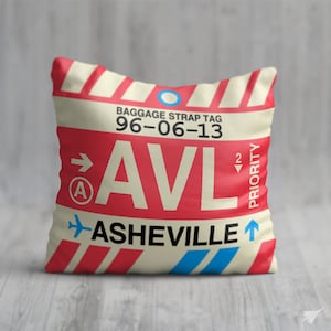 May include: A cream-coloured pillow with a red and white striped pattern. The pillow features a luggage tag with the text "BAGGAGE STRAP TAG 96-06-13 AVL ASHEVILLE" and a blue airplane icon. The luggage tag also has a red arrow pointing to the right with the letter "A" inside a circle and a red arrow pointing down with the number "2" inside a circle. The text "PRIORITY" is written vertically on the right side of the luggage tag.