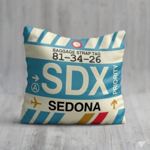 May include: A white and blue throw pillow with a luggage tag design. The tag has the text "Baggage Strap Tag 81-34-26" and "SDX SEDONA" with an arrow pointing to the right. The tag also has a priority label and a circle with the letter "A" inside.