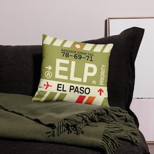 May include: A decorative cushion featuring a luggage tag design. The tag is yellow and white with the text "BAGGAGE STRAP TAG 78-69-71 ELP EL PASO" and a red and orange stripe.