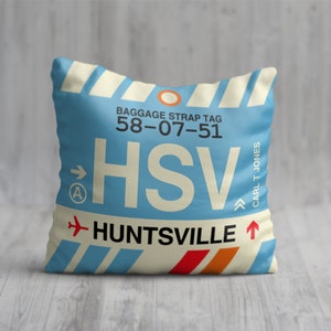 May include: A blue and white pillow with a luggage tag design. The tag has the text "BAGGAGE STRAP TAG 58-07-51 HSV HUNTSVILLE" and an arrow pointing to the right. The tag also has a small airplane icon and an arrow pointing upwards.