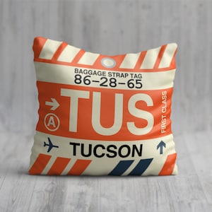 May include: A white and orange luggage tag with the code TUS, the city name Tucson, and the baggage strap tag number 86-28-65. The tag has a red arrow pointing to the right and a letter A inside a circle. The tag also has a small airplane icon and an arrow pointing upwards.
