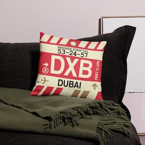 May include: A white and red pillow with a luggage tag design. The tag says "Baggage Strap Tag 53-24-57 DXB DUBAI" with an arrow pointing to the right and a small airplane icon. The tag also says "First Class" with an arrow pointing up.